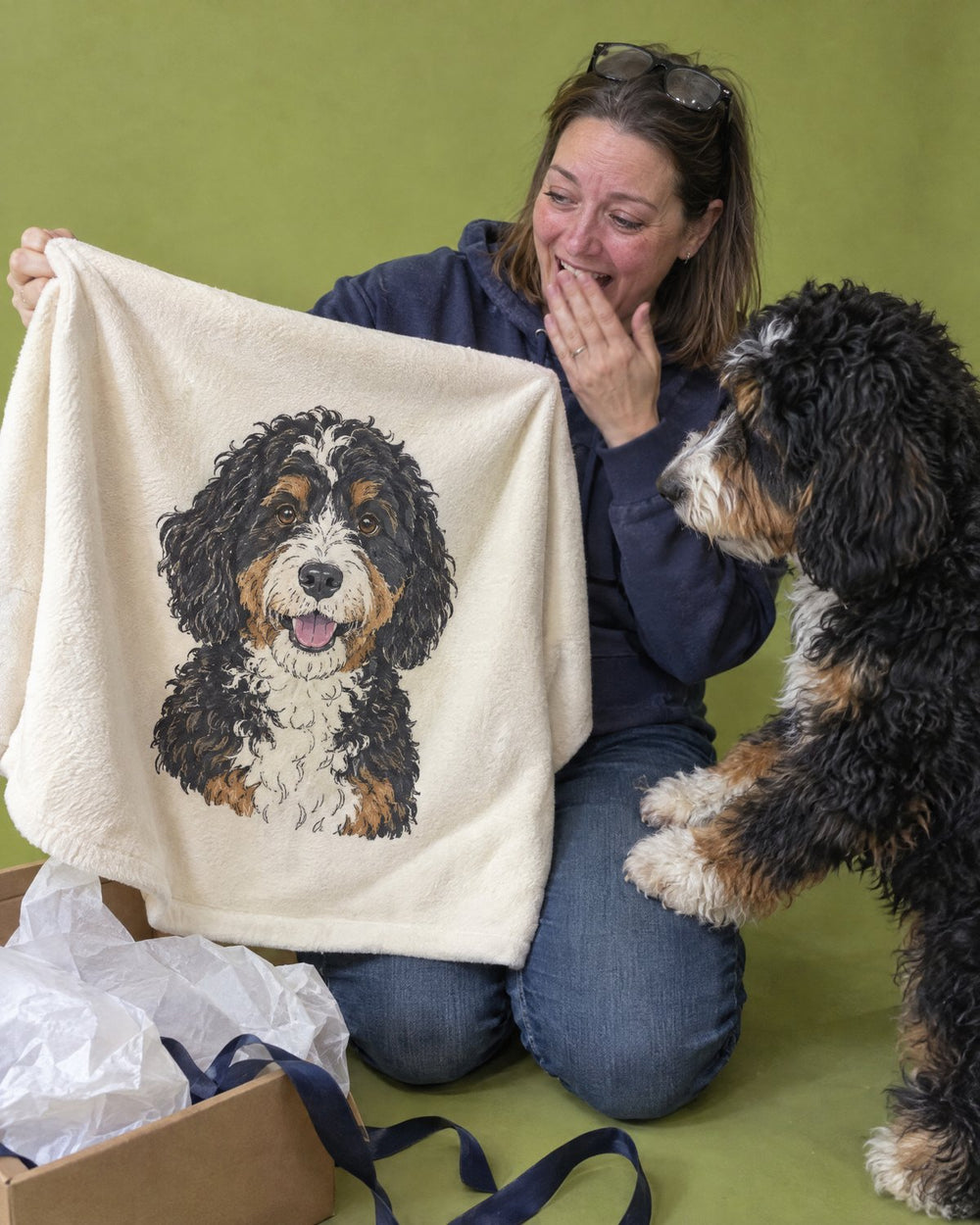 A woman laughing as she unwraps a blanket printed with her Bernedoodle, while the dog looks on